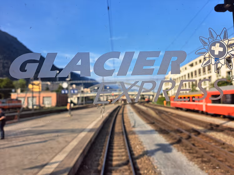Zugfenster des Glacier Express mit Schriftzug vor den Gleisen