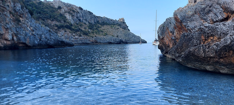 acqua limpida in una baia tra gli scogli, uno yacht in lontananza