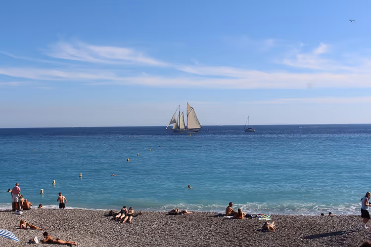 Denizde yüzücüler ve bir yelkenli ile Nice çakıl plajı
