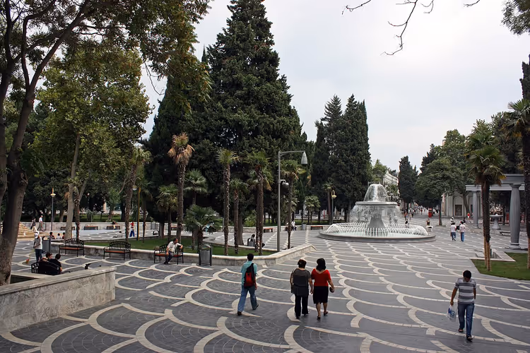 Ağaçlar arasında çeşmeli bir meydanda yürüyen insanlar