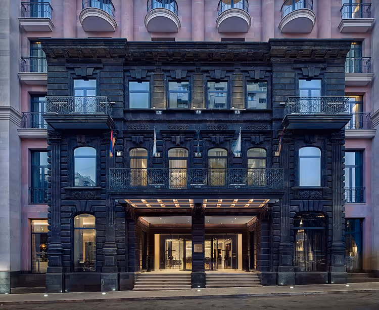 Alexander Hotel'in aydınlatılmış girişli koyu taş cephesi