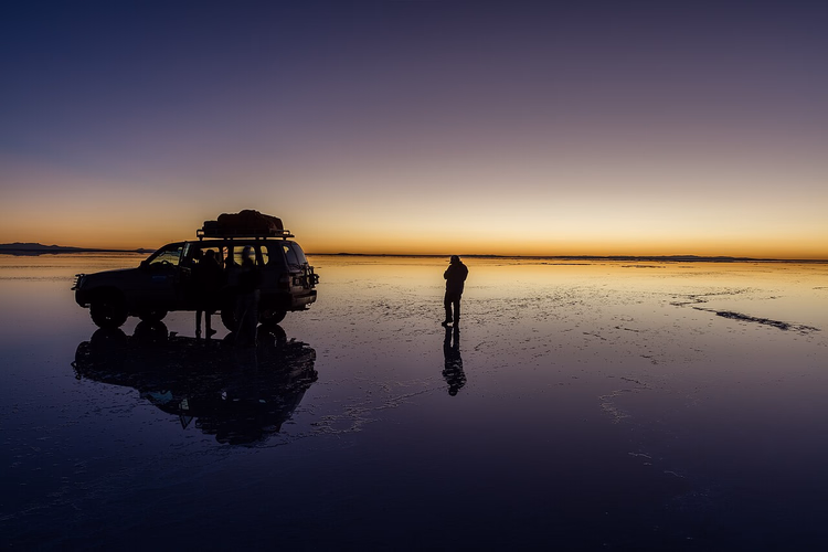 Jeep og reisende speilet i en saltflate ved solnedgang
