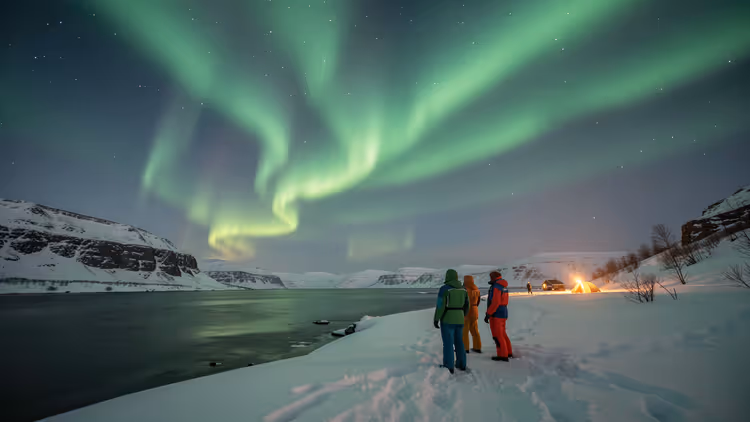 Drie mensen in winterjassen op een besneeuwde fjordoever kijken naar groen noorderlicht, met een kampvuur in de verte