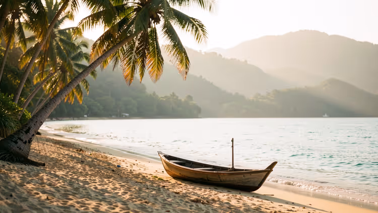Hölzernes Fischerboot am Sandstrand unter schiefen Palmen, dunstige grüne Hügel jenseits einer ruhigen Bucht in Sri Lanka