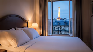 Hotelbett mit weißer Bettwäsche am offenen Balkonfenster mit Blick auf den beleuchteten Eiffelturm über den Dächern von Paris in der Dämmerung
