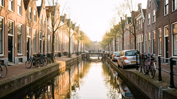 Schmale holländische Gracht mit Backstein-Giebelhäusern, abgestellten Fahrrädern und Autos, kleine Brücke im sanften Morgenlicht