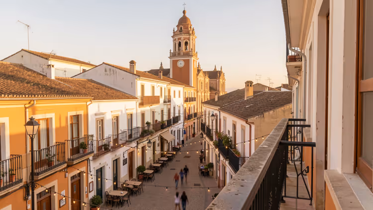 Fußgängerstraße mit Cafétischen, weißen und ockerfarbenen Häusern und barockem Glockenturm bei Sonnenuntergang in Málaga
