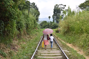 Frau und Kind gehen unter einem rosa Schirm an einem Bahngleis entlang, umgeben von üppigem Grün im Hochland von Sri Lanka