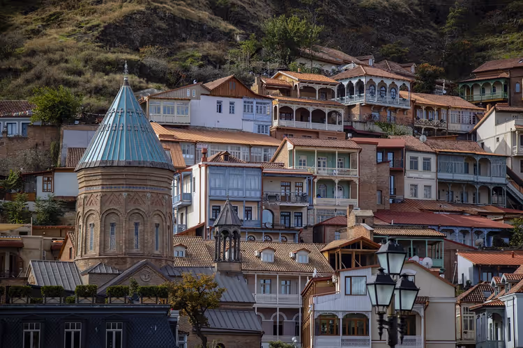 Alte Häuser in Tiflis mit geschnitzten Holzbalkonen an einem Hang neben einer Backsteinkirche mit Kegelkuppel