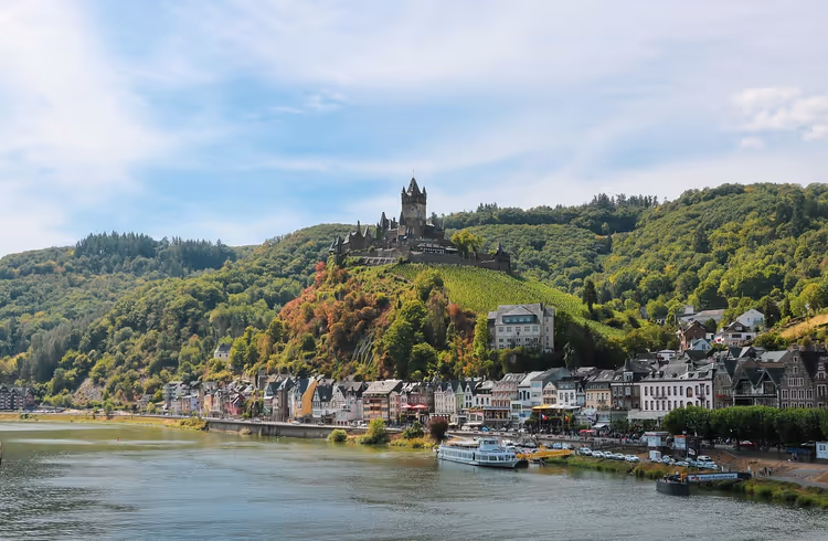 Burg auf einem Hügel über einer Stadt am Moselufer, mit Weinberg am Hang und einem Schiff am Kai