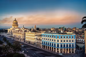 Blau-weißes koloniales Hotelgebäude an einem Boulevard in Havanna, dahinter die goldene Kuppel des Kapitols bei Sonnenuntergang