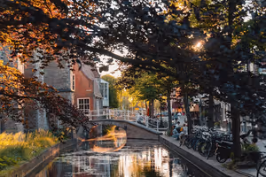 Kleine weiße Bogenbrücke über eine baumgesäumte niederländische Gracht bei Sonnenuntergang, mit abgestellten Fahrrädern am Kai