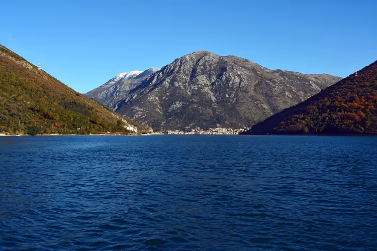 Tiefblaues Wasser der Bucht von Kotor zwischen steilen Hängen, mit einem Städtchen am Fuß eines felsigen, leicht verschneiten Berges