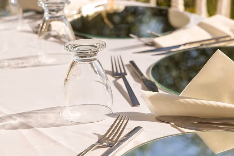 Gedeckter Restauranttisch mit umgedrehten Gläsern, Gabeln, Messern und gefalteten Servietten auf weißem Tischtuch im Sonnenlicht