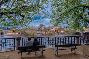 Paar auf einer Bank unter Frühlingsbäumen an der Moldau in Prag, mit Blick über den Fluss zur Prager Burg