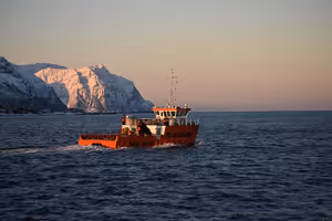 Orange arbetsbåt korsar en mörk fjord nära Tromsø med snötäckta berg som skimrar rosa i skymningen
