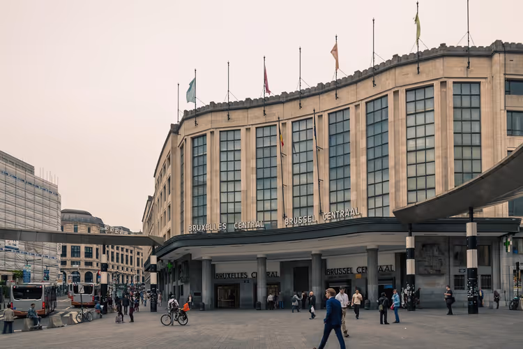 Böjd stenfasad på Bryssels centralstation med flaggor på taket och fotgängare som korsar torget