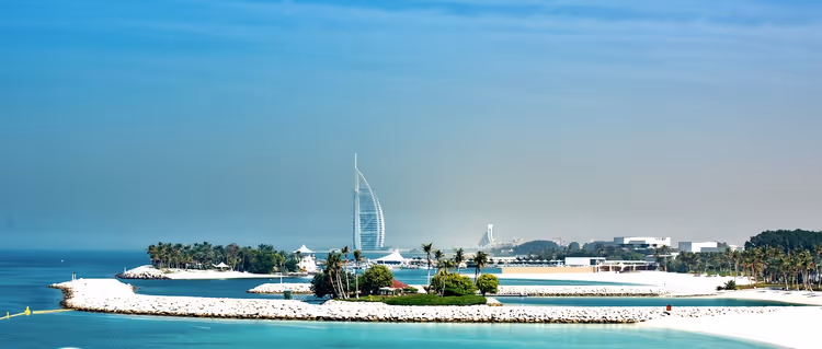 Segelförmiger Hotelturm in Dubai im Dunst hinter einem Wellenbrecher aus Felsen, Palmen und türkisfarbenem Wasser