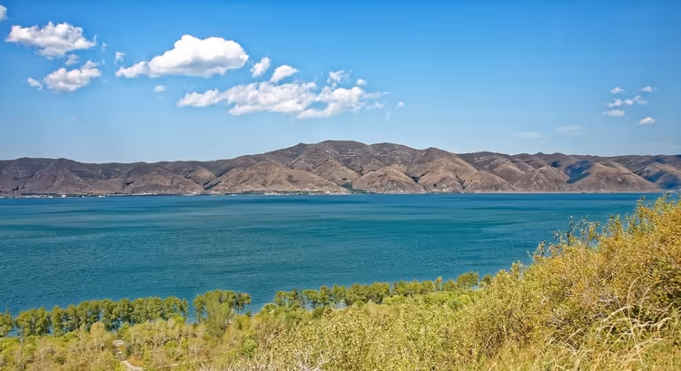 Türkisfarbenes Wasser des Sewansees mit kahlen braunen Hügeln am anderen Ufer und grünen Sträuchern im Vordergrund