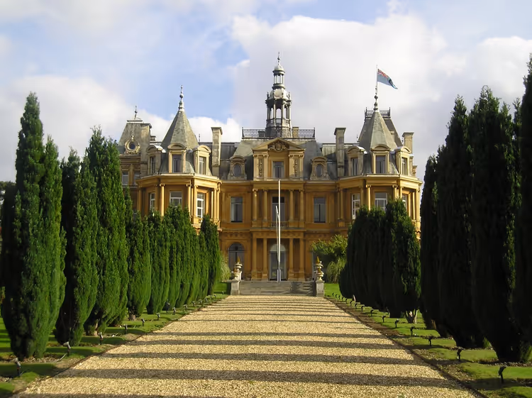 Allée de gravier bordée de hauts cyprès menant à un manoir en pierre dorée aux allures de château, en Grande-Bretagne