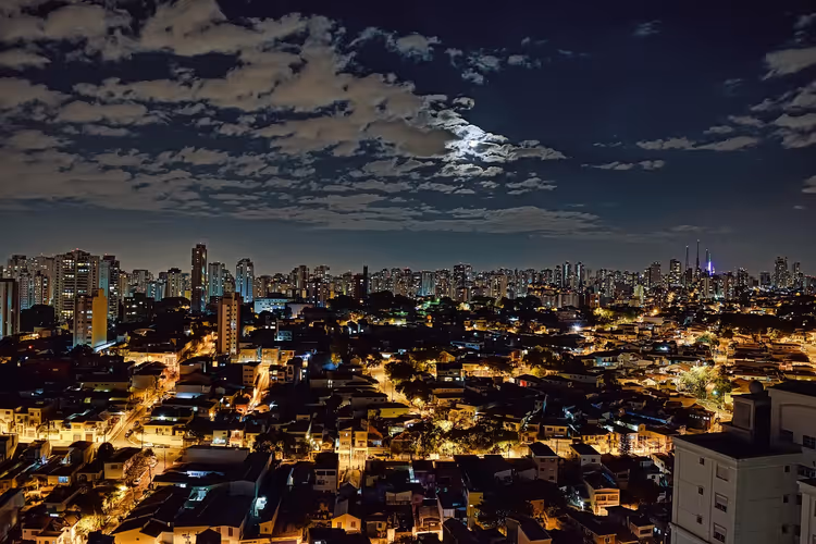 Nächtliche Skyline von São Paulo mit beleuchteten Straßen niedriger Häuser, fernen Hochhäusern und dem Mond hinter Wolken