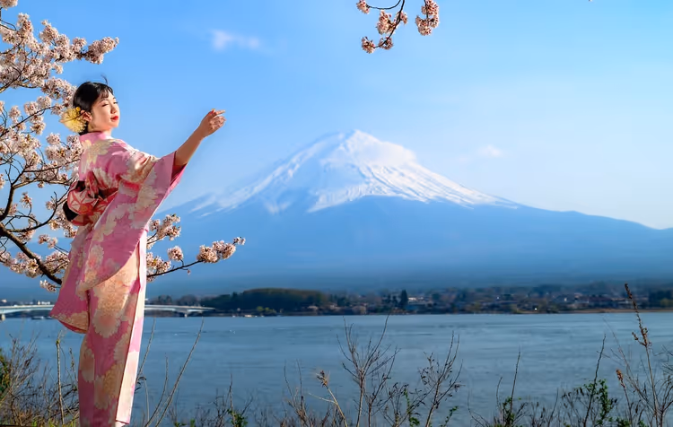 Frau im rosa Kimono greift nach Kirschblüten an einem See, dahinter der schneebedeckte Fuji, Japan