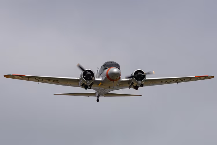 Avião bimotor a hélice antigo, de fuselagem prateada, a voar de frente contra um céu cinzento e encoberto