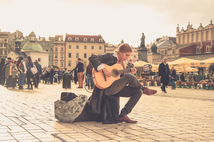 Straßenmusiker kniet mit Akustikgitarre auf einem gepflasterten Altstadtplatz, dahinter Menschenmenge und Marktschirme