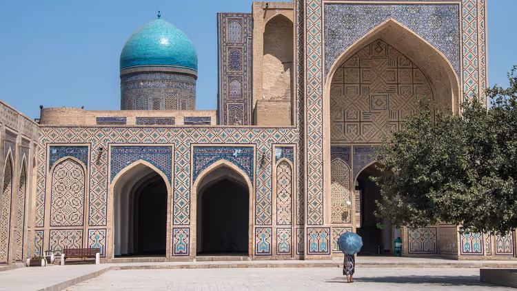 Gekachelte Fassade einer Medrese mit Spitzbögen und türkisfarbener Kuppel in Usbekistan, eine Frau mit blauem Schirm geht vorbei