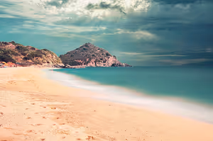 Leerer Sandstrand in Italien mit türkisfarbenem Meer, felsiger Landzunge und Sonnenstrahlen, die durch schwere Wolken brechen