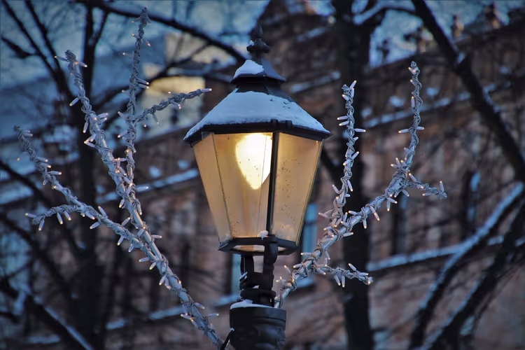 Verschneite Straßenlaterne leuchtet zwischen Zweigen mit Lichterketten an einem Winterabend in Helsinki
