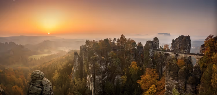 Basteibrücke zwischen Sandsteinfelsen und Herbstwald in der Sächsischen Schweiz bei Sonnenaufgang über einem nebligen Tal