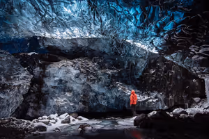Person in orangefarbener Jacke steht in einer blauen Eishöhle unter der gewellten Decke des Gletschers Vatnajökull