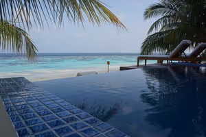 Infinity-Pool mit blauem Fliesenrand vor einer türkisfarbenen Lagune auf den Malediven, Sonnenliegen unter Palmen