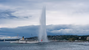 Fontäne Jet d'Eau über dem Genfersee, davor ein weißer Raddampfer unter schweren grauen Wolken