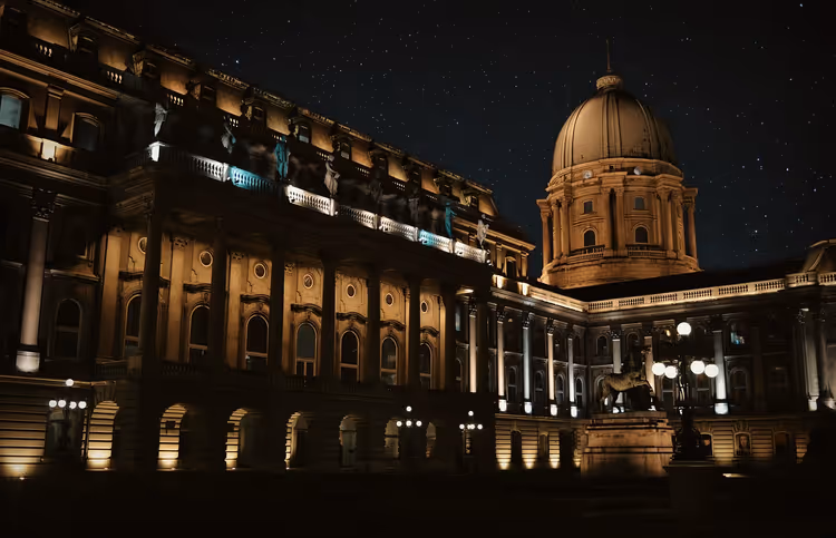 Innenhof des Burgpalasts in Budapest bei Nacht, angestrahlte Fassade, Arkaden und grüne Kuppel unter Sternenhimmel