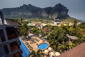 Hotelpools mit Liegen und Sonnenschirmen zwischen Palmen, dahinter steile Kalksteinfelsen über einem Ferienort in Thailand