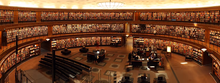 Sala circolare di una biblioteca con tre ordini di scaffali curvi lungo le pareti e tavoli di lettura al piano sottostante