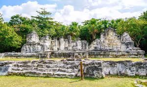 Verwitterte steinerne Maya-Ruinen mit Säulenreihen auf einem grasbewachsenen, von Palmen umgebenen Gelände in Cancún