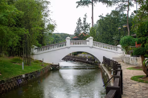 Weiße Bogenbrücke für Fußgänger mit Balustraden über einem stillen Kanal in einem üppigen tropischen Park, daneben ein Steinweg