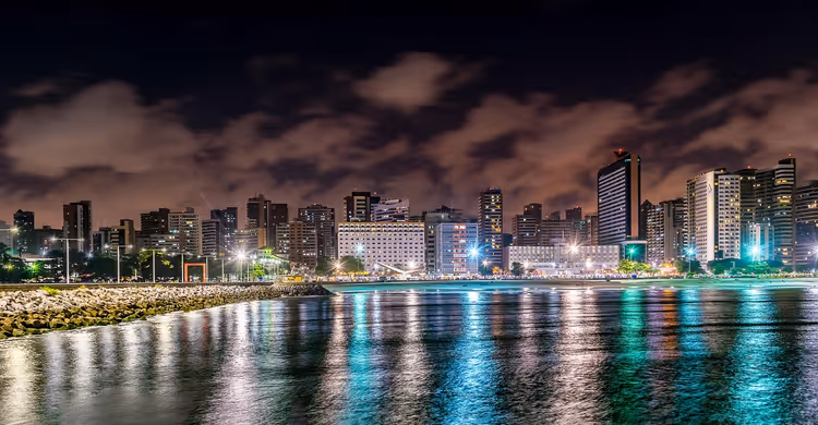 Skyline von Fortaleza bei Nacht mit erleuchteten Hochhäusern und bunten Spiegelungen im Wasser neben einem steinernen Wellenbrecher