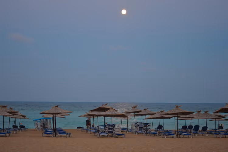 Vollmond über ruhigem Meer und Reihen strohgedeckter Sonnenschirme mit blauen Liegen an einem leeren bulgarischen Strand in der Dämmerung