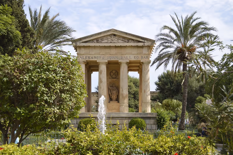 Klassizistisches Tempelmonument aus Stein mit Säulen hinter einem Brunnen, eingerahmt von Palmen und Sträuchern in einem Garten auf Malta