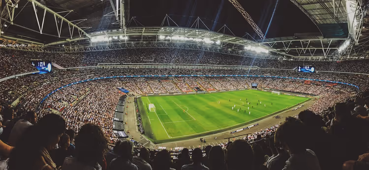 Vol voetbalstadion bij avond, gezien vanaf de bovenste tribune, met schijnwerpers die het groene veld verlichten tijdens een wedstrijd