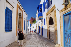 Kopfsteinpflastergasse in Tunesien mit weiß getünchten Häusern, blauen Fensterläden und Türen und fotografierenden Touristen