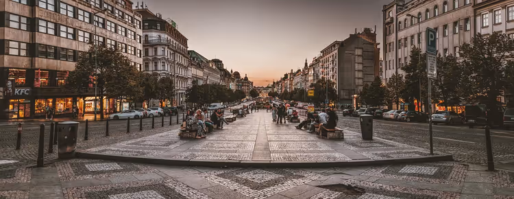 Wenzelsplatz in Prag in der Dämmerung, Menschen auf Bänken am gepflasterten Mittelstreifen zwischen erleuchteten Schaufenstern