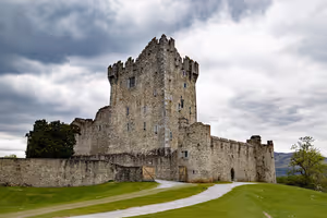 Steinerner Wohnturm und Zinnenmauern von Ross Castle in Killarney unter schweren grauen Wolken, ein Weg führt über den Rasen