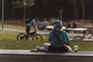 Frau mit Kopftuch sitzt mit einem Eisgetränk auf einer Parkbank, hinter ihr fährt ein Radfahrer auf dem Weg vorbei