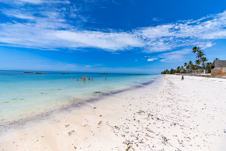Breiter weißer Sandstrand auf Sansibar mit flachem türkisfarbenem Wasser, einigen Badenden, Holzbooten und Palmen unter blauem Himmel