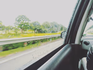 Regentropfen auf dem Seitenfenster eines Autos, dahinter unscharf eine Leitplanke, grüne Bäume und ein grau verhangener Himmel